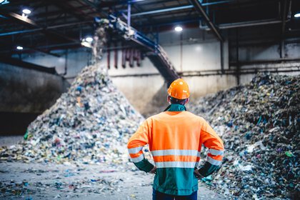 Eine Person in Schutzkleidung und Helm steht in einer Recycling- oder Abfallanlage vor großen Haufen sortiertem Müll und beobachtet den laufenden Prozess.