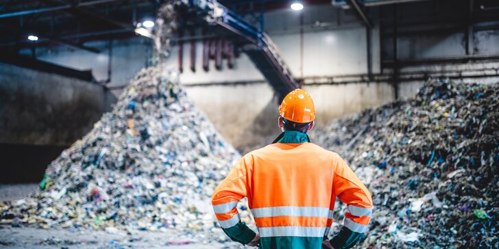 Eine Person in Schutzkleidung und Helm steht in einer Recycling- oder Abfallanlage vor großen Haufen sortiertem Müll und beobachtet den laufenden Prozess.