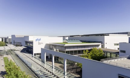 Modern building complex with elegant design, surrounded by trees and streets, under a blue sky. The Messe München logo can be seen on one building.