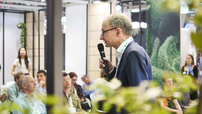 A man speaks into a microphone on an exhibition stage, surrounded by seated spectators and green plants.