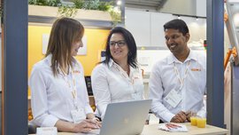 Three exhibitors in white shirts smile and chat, a woman holds a laptop in her hand in an industrial plant.