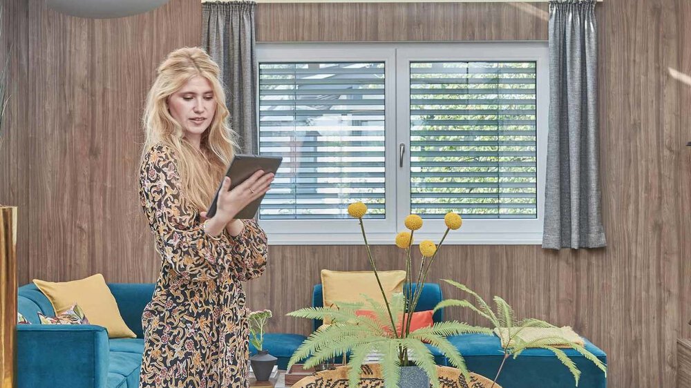 Eine Frau in einem braun gemusterten Kleid steht mit einem Tablet in der Hand in einem gemütlichen Raum mit Esstisch, großem Fenster und bunten Kissen.