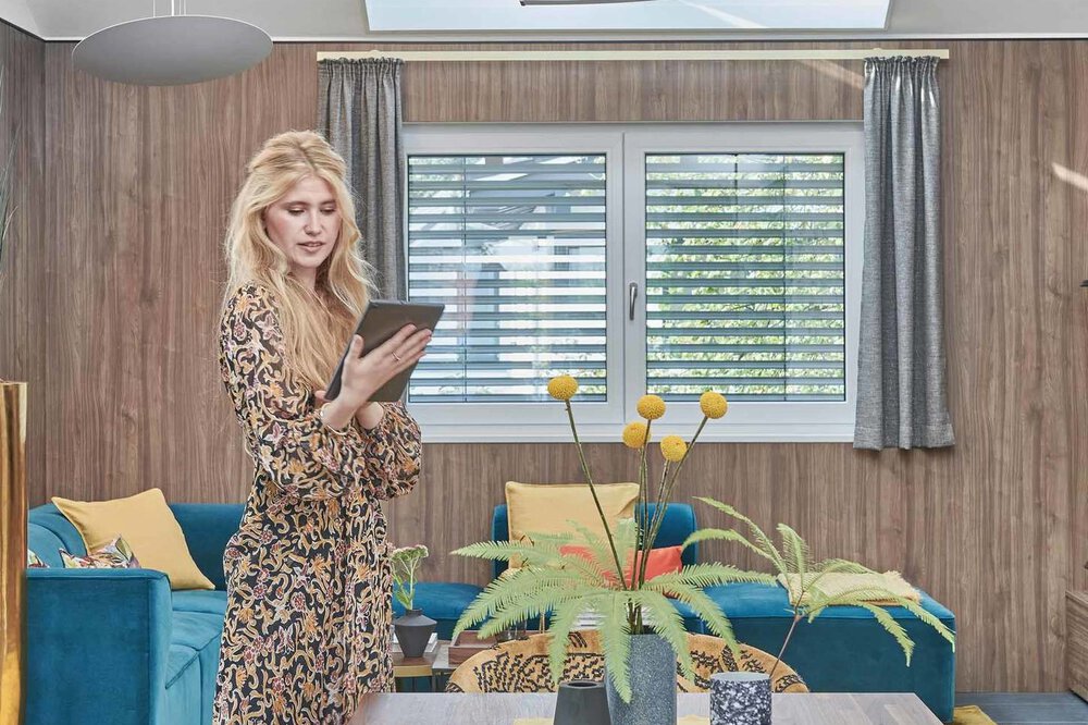 Eine Frau in einem braun gemusterten Kleid steht mit einem Tablet in der Hand in einem gemütlichen Raum mit Esstisch, großem Fenster und bunten Kissen.