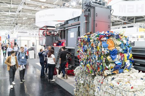 Besucher an einem Messestand betrachten eine Ballenpressmaschine mit 3 davor gestapelten Ballen aus verschiedenen Wertstoffen.