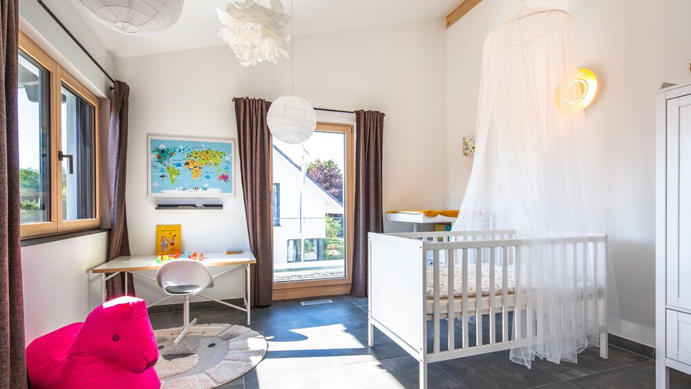 Helles Kinderzimmer mit Kinderbett, rundem Teppich, rosa Sitzsack, Schreibtisch mit Weltkarte und großen Fenstern mit braunen Vorhängen.