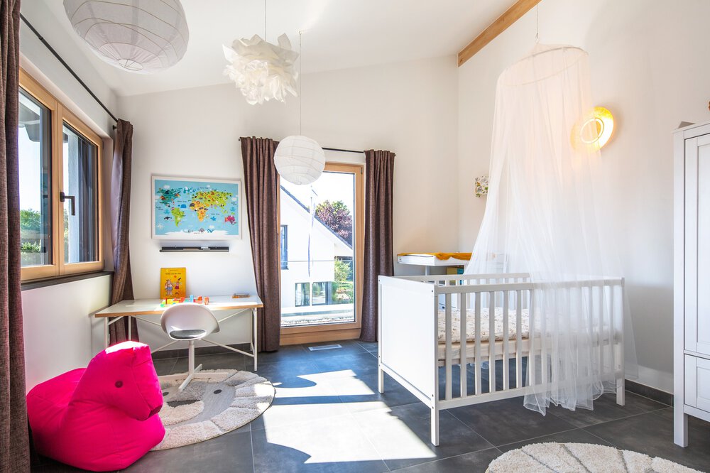 Helles Kinderzimmer mit Kinderbett, rundem Teppich, rosa Sitzsack, Schreibtisch mit Weltkarte und großen Fenstern mit braunen Vorhängen.