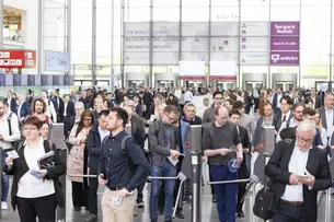 Entrance to the analytica trade fair with many people going through the turnstiles. In the background hangs an analytica poster with 