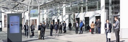 Menschen stehen unter sonnigem Himmel vor dem Messeeingang und unterhalten sich. Im Vordergrund ist eine Willkommenstafel der LOPEC.
