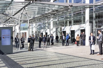 People stand under a sunny sky in front of the trade fair entrance and chat. In the foreground is a LOPEC welcome board.