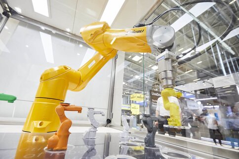 Gelber Roboterarm platziert an einem Messestand farbige Robotermodelle in Aussparungen in einer Glasvitrine.