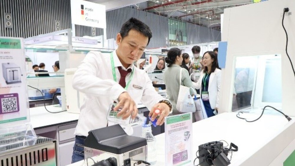 A man in a white shirt demonstrates laboratory equipment at a busy stand at a technology trade fair.