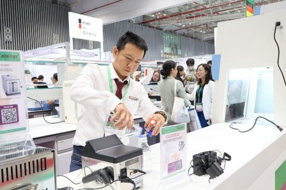 A man in a white shirt demonstrates laboratory equipment at a busy stand at a technology trade fair.