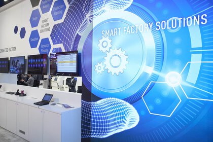 Ausstellerstand mit einer abstrakt verzierten blauen Wand, auf der „Smart Factory Solutions“ steht sowie mehreren Monitoren und einem Laptop auf einem langen Tresen.