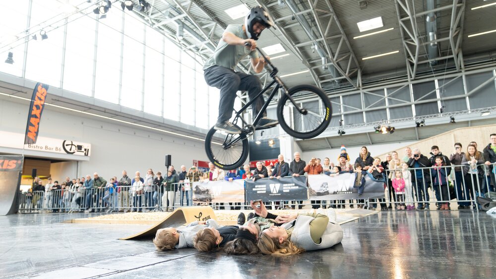 Ein BMX-Fahrer springt über mehrere am Boden liegende Personen während einer Show in der Messehalle. Hinter einer Absperrung beobachten zahlreiche Zuschauer die Aktion.
