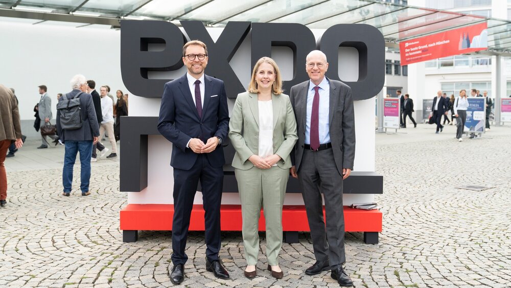 Three people in business attire are standing in front of a large EXPO REAL logo in an outdoor area, surrounded by trade fair visitors at the exhibition grounds.