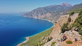 Blick über eine blaue Bucht auf Kreta. Auf einer Aussichtsplattform, an einem der teilweise bewachsenen Berghänge, steht eine Gruppe Menschen und blickt hinunter.