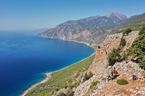 Blick über eine blaue Bucht auf Kreta. Auf einer Aussichtsplattform, an einem der teilweise bewachsenen Berghänge, steht eine Gruppe Menschen und blickt hinunter.