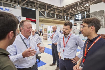 Five men discussing at a trade fair, surrounded by stands and displays in a large hall.