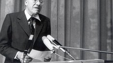 A black and white photograph of an elderly gentleman standing behind a lectern with microphones and reciting something. The sign 