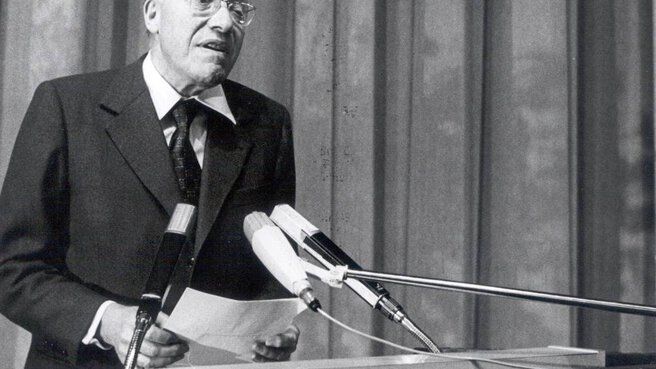 Eine schwarz-weiß Aufnahmen auf der ein älterer Herr hinter einem Pult mit Mikrophonen steht und etwas vorträgt. Der Schirftzug 