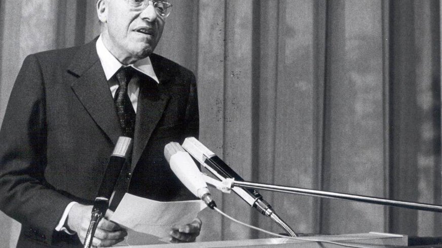 A black and white photograph of an elderly gentleman standing behind a lectern with microphones and reciting something. The sign 
