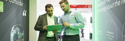 An exhibitor shows a visitor something on a tablet. The two men are standing between two large display walls.
