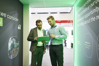 An exhibitor shows a visitor something on a tablet. The two men are standing between two large display walls.