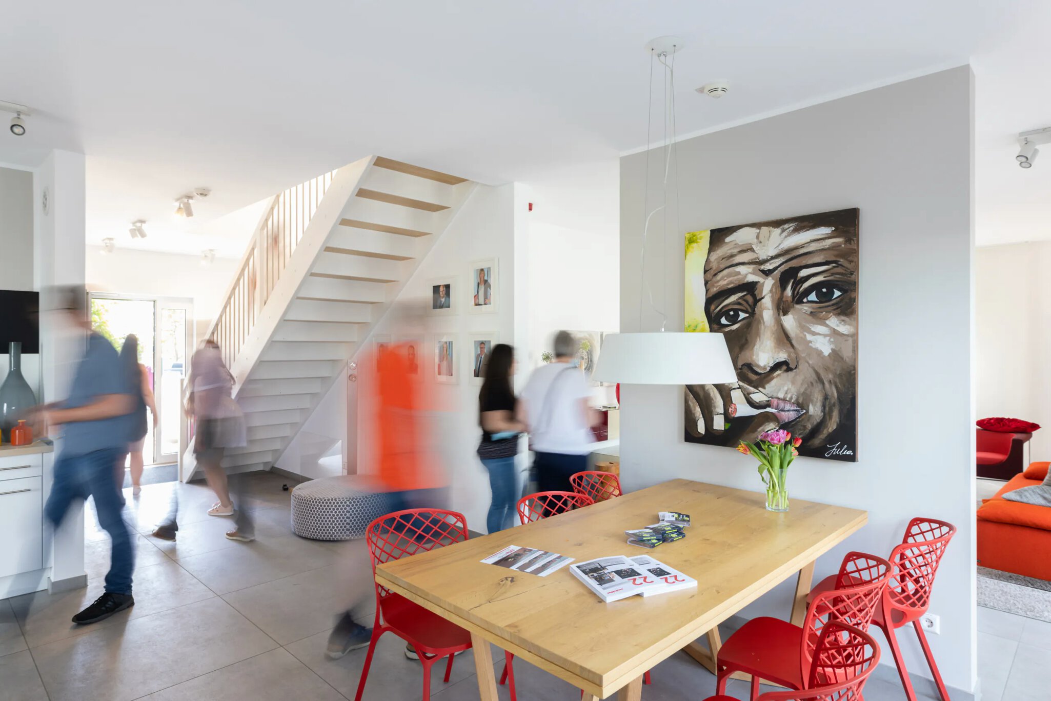 Ein großer offener Wohnbereich mit einem Tisch und Stühlen, Sofas, Küche und einer Treppe ins Obergeschoss und den Schatten von sich bewegenden Besuchern.