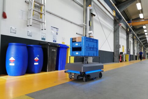 Autonom fahrende Palette befördert zwei blaue Kisten durch eine Lagerhalle.
