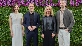 Vier Personen stehen vor einer grünen Wand mit lila Blumen und lächeln in die Kamera.