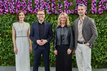 Four people stand in front of a green wall with purple flowers and smile at the camera.