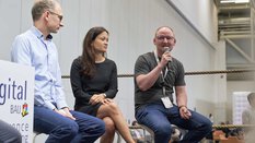 Three people are sitting in a panel discussion. The barrier of a boxing ring can be seen behind them.