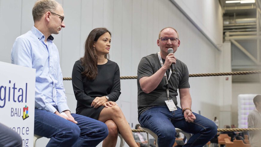 Three people are sitting in a panel discussion. The barrier of a boxing ring can be seen behind them.