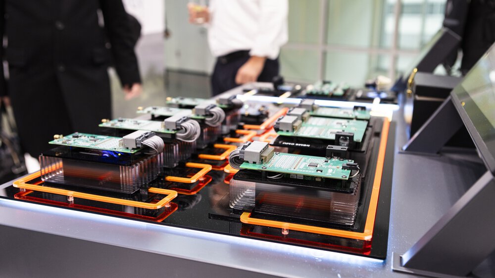 Elektronische Leiterplatten mit Kabeln und Kühlkörpern liegen in mehreren Reihen als Exponat auf einem Messetisch im Vordergrund sind die Bauteile zu sehen im Hintergrund stehen Personen