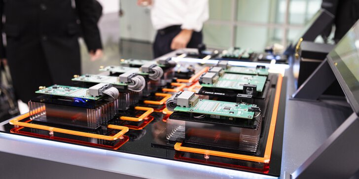 Electronic circuit boards with cables and heat sinks are arranged in several rows as an exhibit on a trade fair table in the foreground the components are visible in the background people are standing