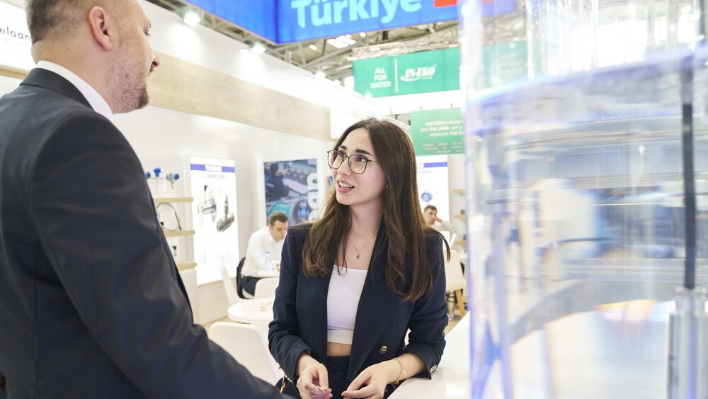Zwei Geschäftsleute, die sich angeregt und freundlich unterhalten. Die Dame erklärt dem Herrn die neuesten Technologien ihrer ausstellenden Firma. Im Hintergrund ist das Logo des türkischen Gemeinschaftsstandes zu sehen.