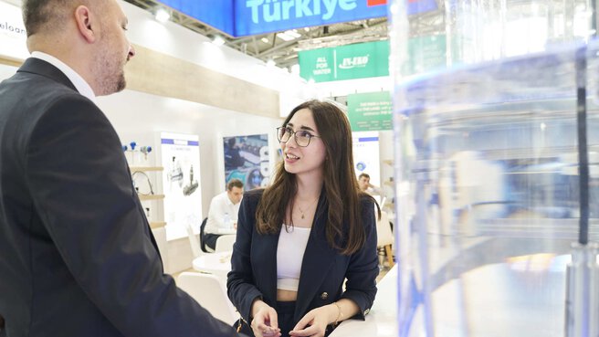 Two business people having a lively and friendly conversation. The lady explains the latest technologies of her exhibiting company to the gentleman. The logo of the Turkish joint pavilion can be seen in the background.