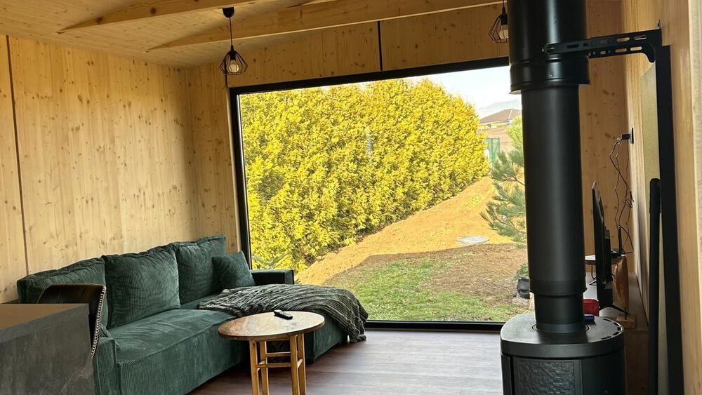 Gemütliches Interieur einer Holzhütte mit grünem Sofa, schwarzem Holzofen und einem großen Fenster mit Blick auf den Garten.