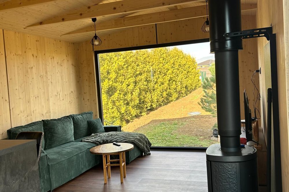 Gemütliches Interieur einer Holzhütte mit grünem Sofa, schwarzem Holzofen und einem großen Fenster mit Blick auf den Garten.