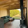 Gemütliches Interieur einer Holzhütte mit grünem Sofa, schwarzem Holzofen und einem großen Fenster mit Blick auf den Garten.