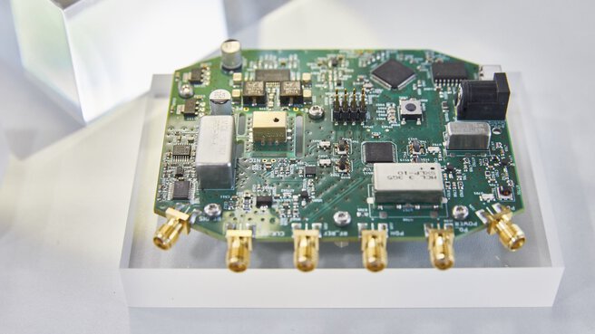 Eine Leiterplatte mit Elektronischen Komponenten und mehreren Analoganschlüssen, präsentiert auf einem Plexiglaswürfel an einem Messestand.