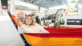 Eine lächelnde Frau entspannt sich in einer Hängematte und hält ein Getränk hoch, während im Hintergrund Wohnmobile zu sehen sind.