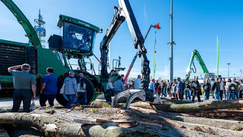 Ein grüner Forstharvester mit einem langen mechanischen Greifarm, der auf einer Fachmesse für Outdoor-Ausrüstung gefällte Baumstämme bearbeitet.
