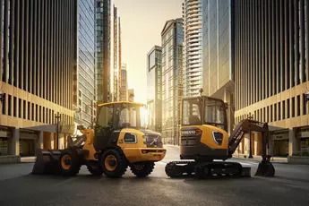 Zwei gelbe Volvo-Baumaschinen stehen bei Sonnenuntergang zwischen hohen Gebäuden.