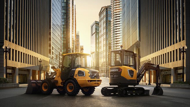 Zwei gelbe Volvo-Baumaschinen stehen bei Sonnenuntergang zwischen hohen Gebäuden.