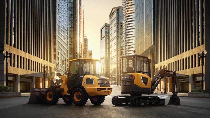 Two yellow Volvo construction machines stand between tall buildings at sunset.