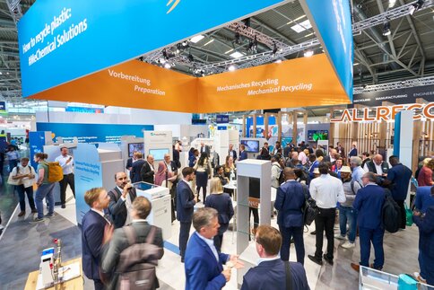 Belebter Messestand zum Thema Kunststoffrecycling auf einer Fachmesse, mit zahlreichen Besucherinnen und Besuchern, Präsentationen zu mechanischem Recycling und Vorbereitungstechnologien sowie verschiedenen Ausstellungsflächen und Informationsdisplays.