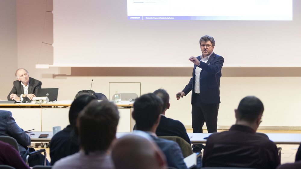 Ein Mann steht auf einer Bühne vor einer Leinwand mit Präsentationsfolie und hält einen Vortrag vor einem interessierten Publikum.