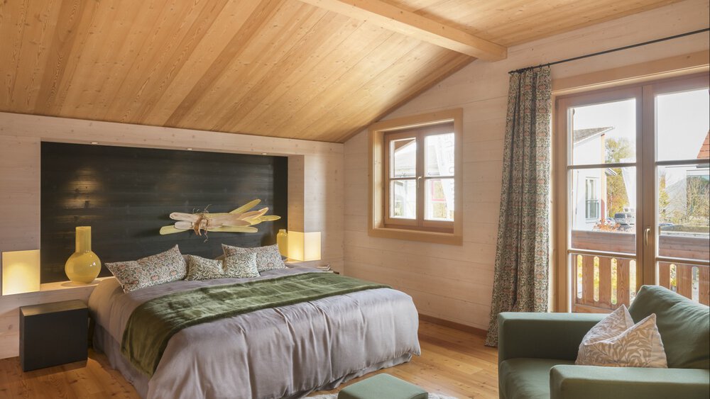 Gemütliches Schlafzimmer mit Holzdecke, großem Bett, Sessel und Fenster, das auf einen Balkon mit malerischer Aussicht führt.