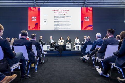 Sechs Personen in Businesskleidung und mit einem Mikrofon sitzen auf einer Bühne der EXPO REAL vor Publikum und sprechen über das „Flexible Housing Forum“.
